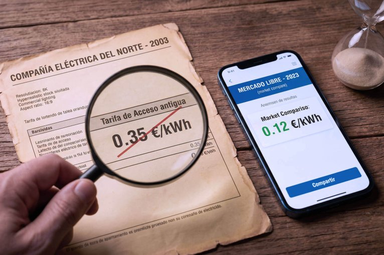 Vista cenital en un escritorio de madera que muestra una factura de luz antigua del 2003 bajo una lupa con un precio de 0.35 euros/kWh tachado en rojo, comparado con un teléfono inteligente moderno al lado que muestra "MERCADO LIBRE - 2023" con un precio verde de 0.12 euros/kWh. Un reloj de arena está en la esquina.