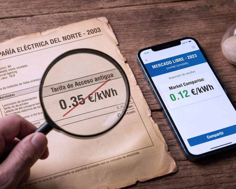 Vista cenital en un escritorio de madera que muestra una factura de luz antigua del 2003 bajo una lupa con un precio de 0.35 euros/kWh tachado en rojo, comparado con un teléfono inteligente moderno al lado que muestra "MERCADO LIBRE - 2023" con un precio verde de 0.12 euros/kWh. Un reloj de arena está en la esquina.